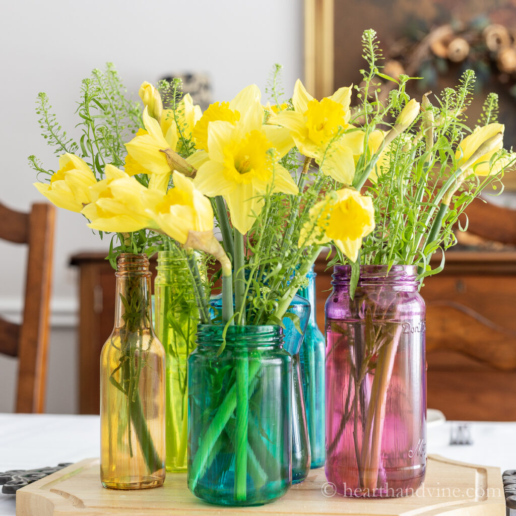 Daffodils and greenery in a group of tinted glass jars creating a pretty centerpiece on the dining room table.