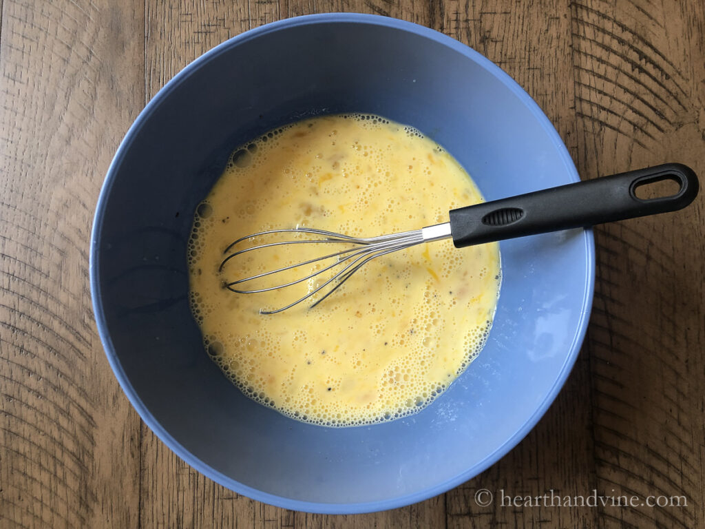 Mixing bowl with eggs and milk and a wire whisk.