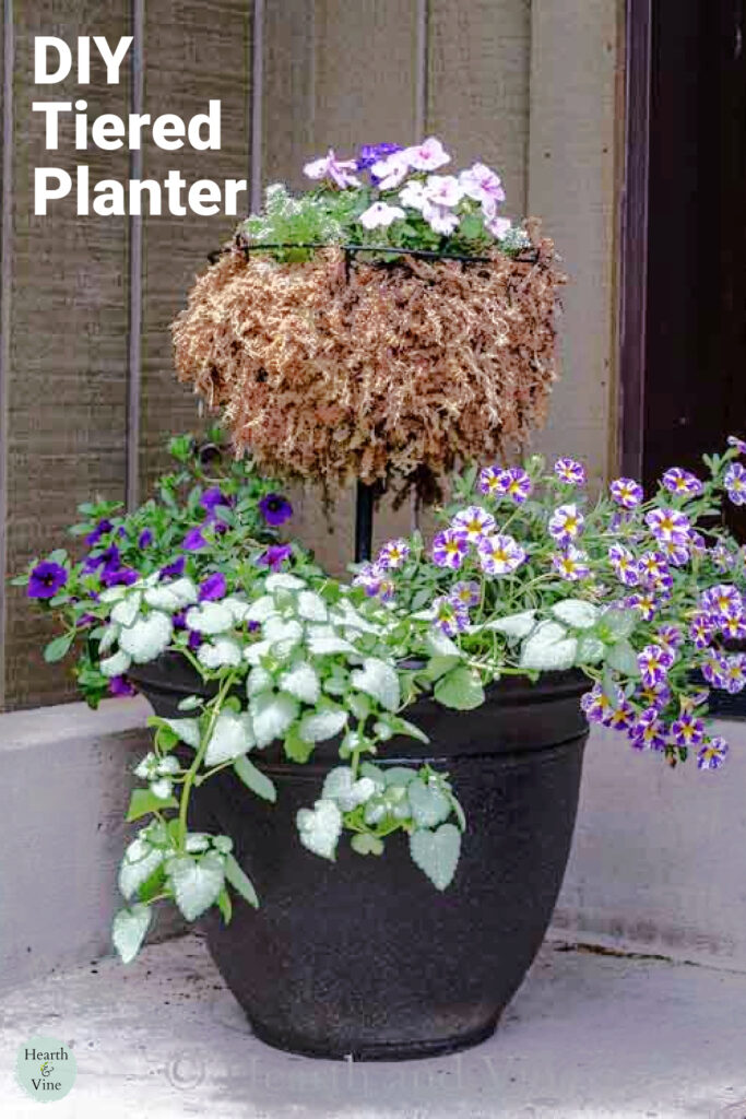 Two tiered flower planter with rounded basket top filled with trailing purple and pink flowers.