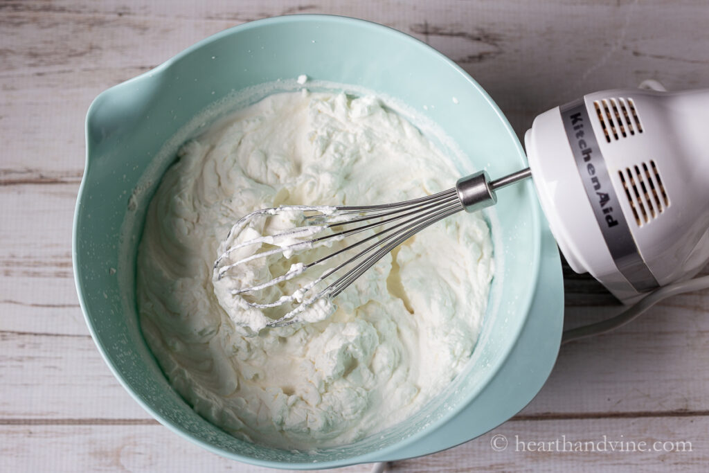 Whipped cream with stiff peaks in a bowl with a hand mixer.