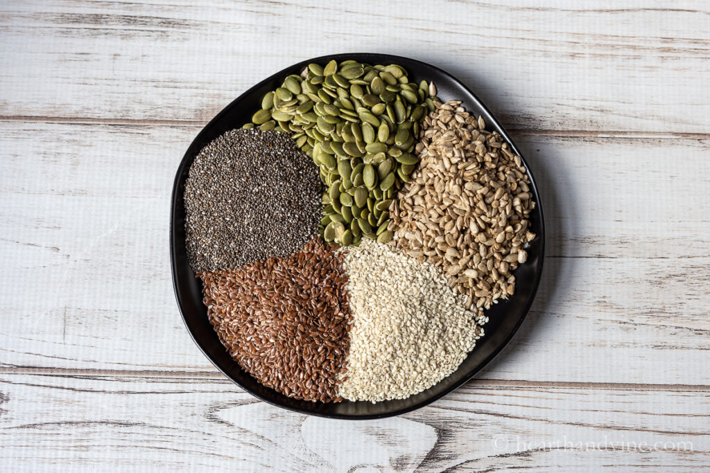 Pumpkin seeds, sunflower seeds, flax seeds, chia seeds and white sesame seeds on a black plate.