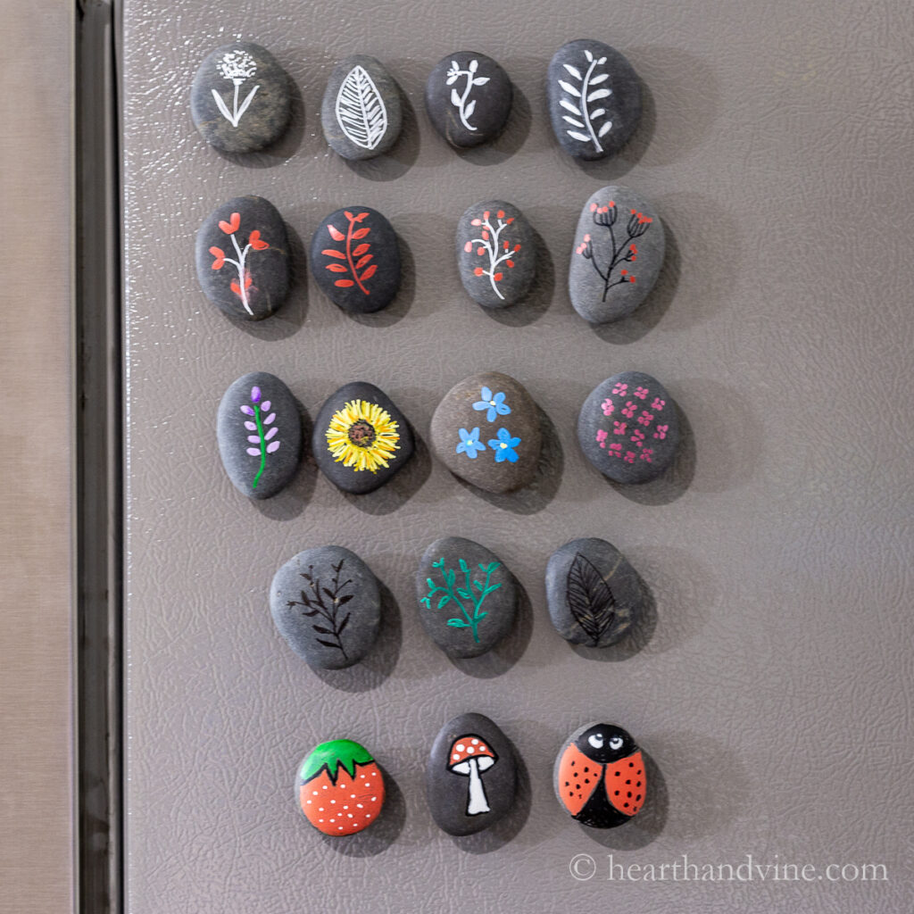 Various painted rock magnets on a refrigerator including  leaves, flowers, a ladybug and a strawberry.