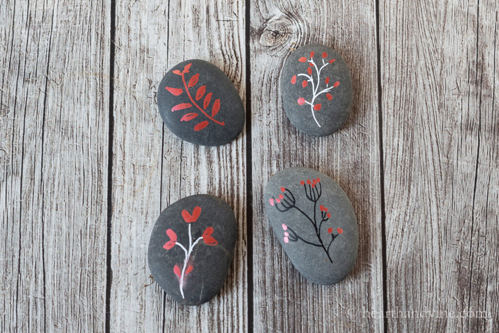 Four painted rocks using white, black and red paint in leaf and branch graphics.