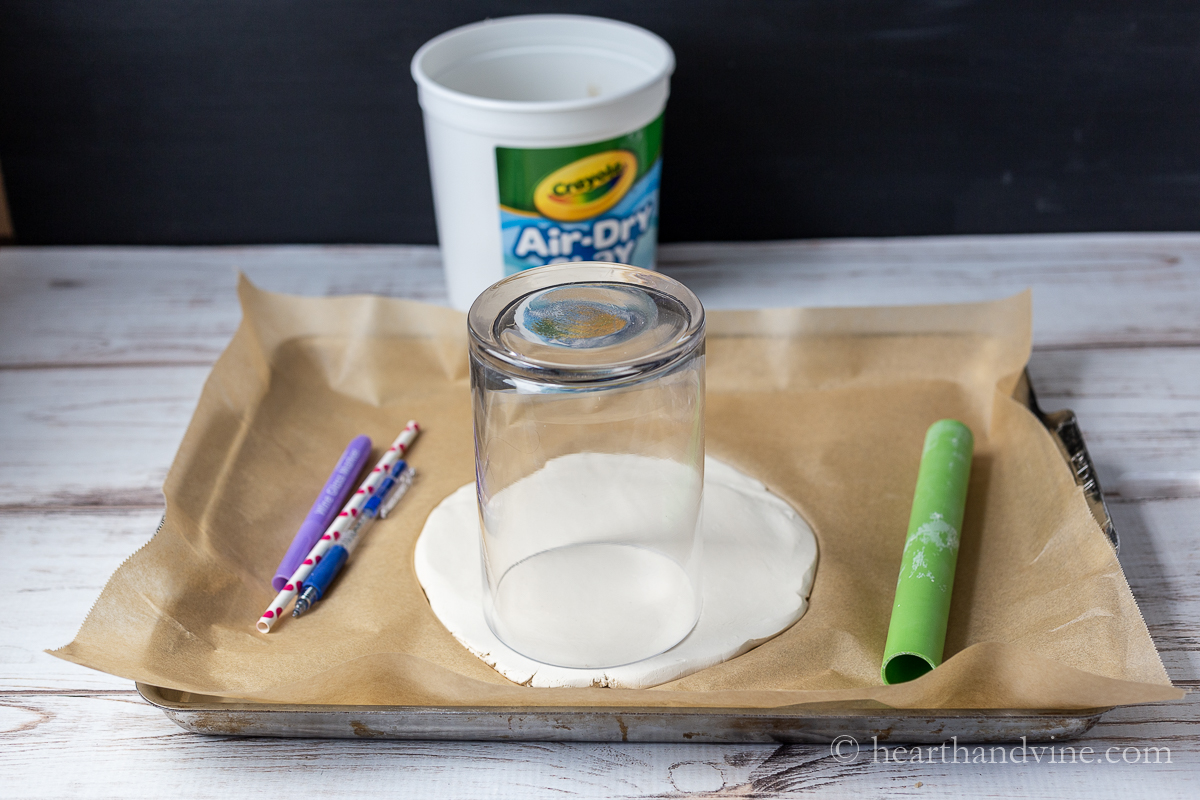 A tray covered with parchment. Rolled out clay with glass cylinder vase upside-down, A marker, pen and straw and a plastic roller.