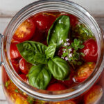 Top view of jar of marinated tomatoes with fresh basil and thyme leaves.