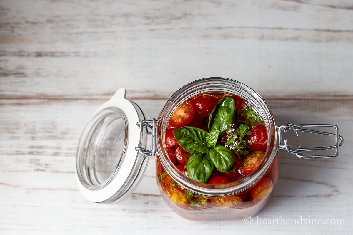 Open jar of marinated tomatoes with some basil leaves and thyme on top.