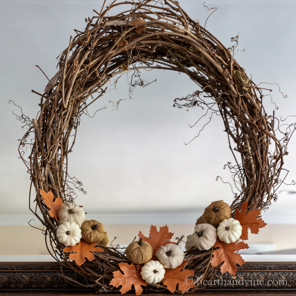 Grapevine wreath with mini pumpkins and faux leather oak leaves leaning on a mantel.