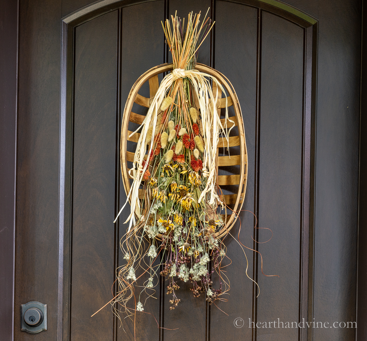 Dried flowers and grasses gathered with raffia and tied to a tobacco basket hanging on a brown door.