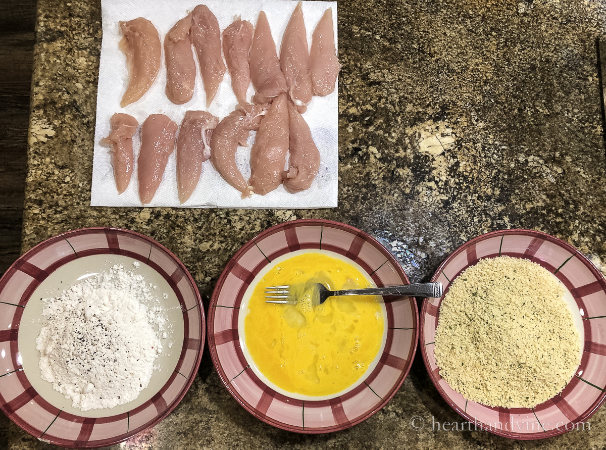Dredging station with raw chicken tenders, a bowl of flour, a bowl of beaten eggs and a bowl of seasoned Panko breadcrumbs.