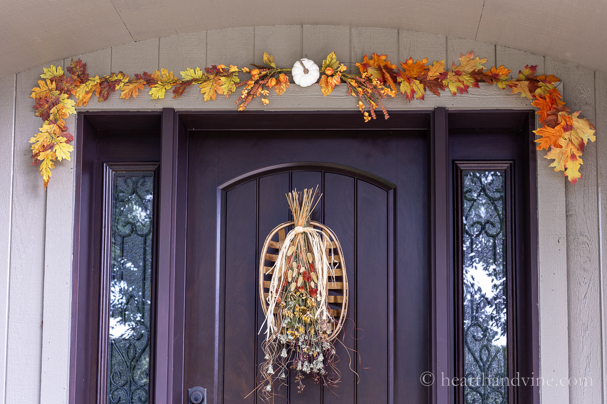 Autumn leaves and a mini white pumpkin garland above a front door with a dried flower and tobacco basket wreath.