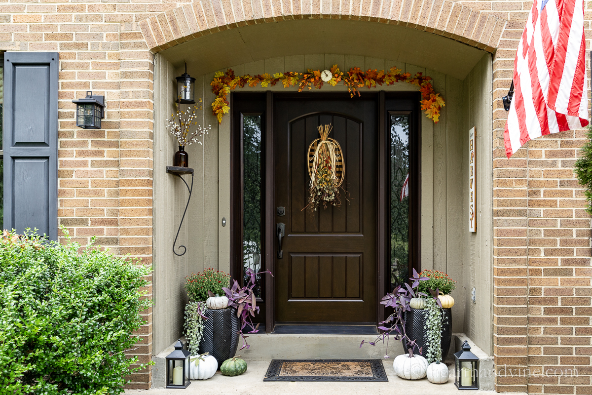 Fall front door decorations including a dried flower fall swag, an artificial leaf garland in autumn colors, faux pumpkins, lanterns and a fall harvest sign.