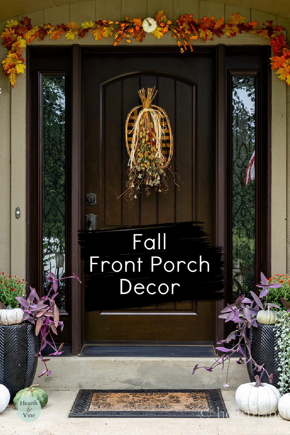Front door decorated for fall with an artificial fall leaf garland, faux pumpkins, a fall swag hanging on the door and orange mums in planters.