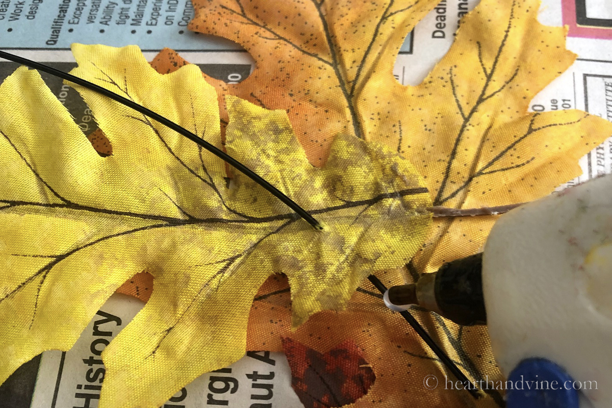 Thick black wire pushed through artificial fall leave. Hot glue gun attaching leaves to wire and several artificial fall leaves strung on a wire.