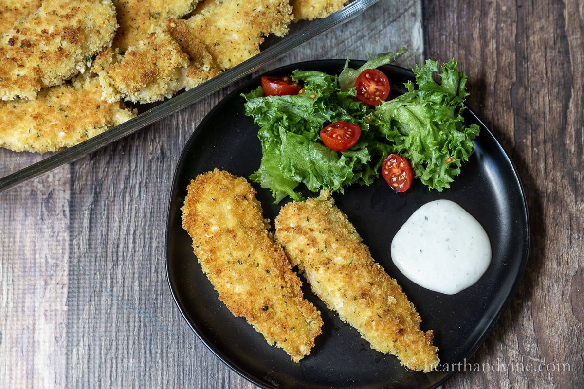 Serving of panko chicken strips with a side of ranch and a salad next to a baking dish with more chicken.