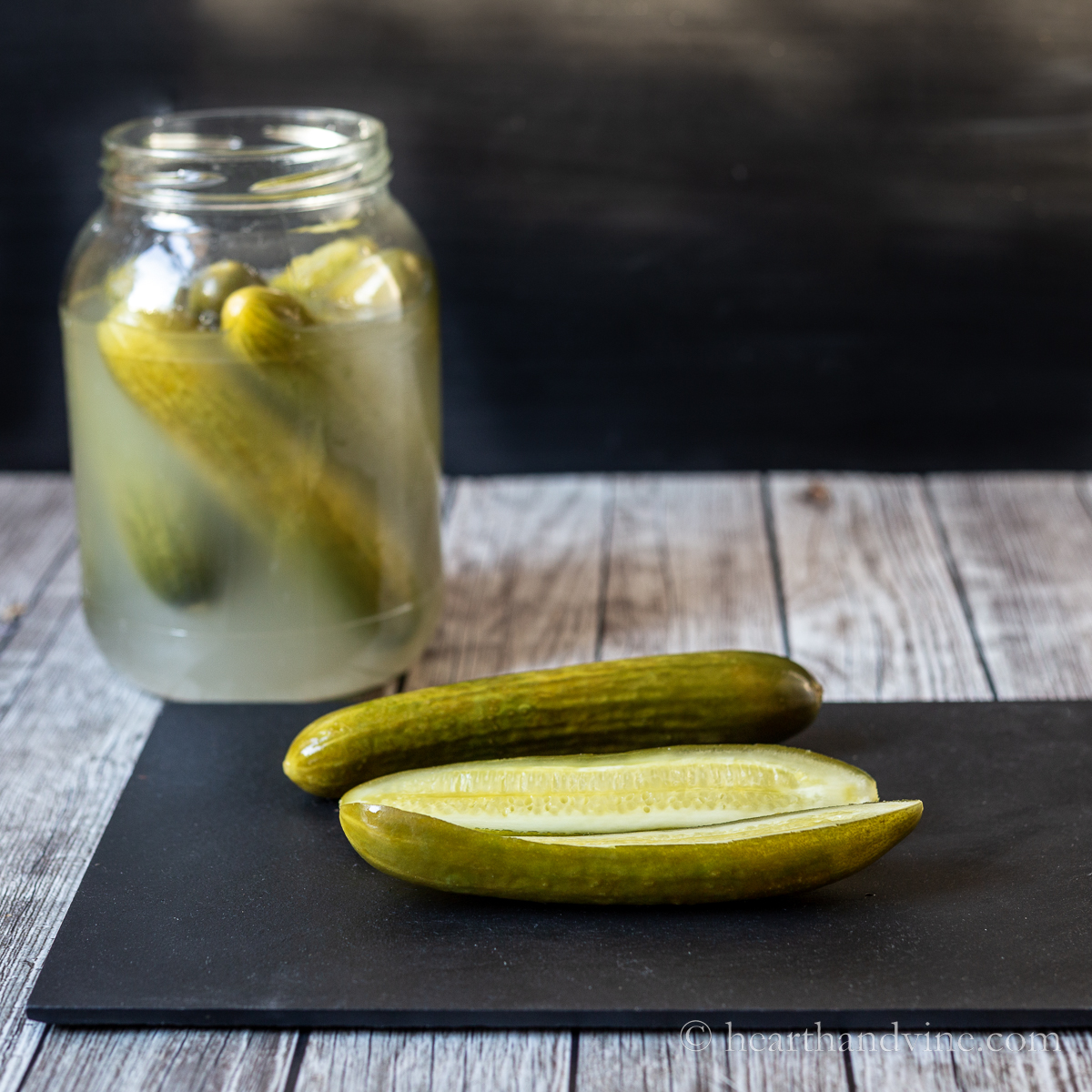 Fermented pickles on a black slate next to a jar of pickles.