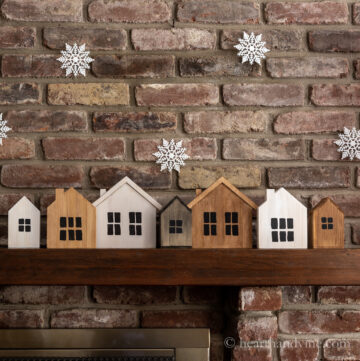 Painted wooden houses on a mantel with snowflakes hanging from above.