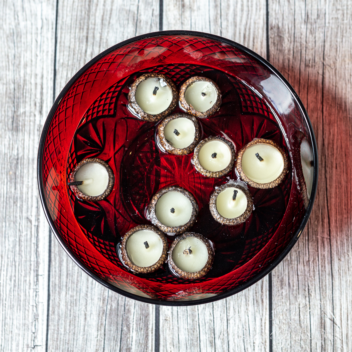 An ornate red bowl filled with water and several beeswax acorn cap candles floating on top.