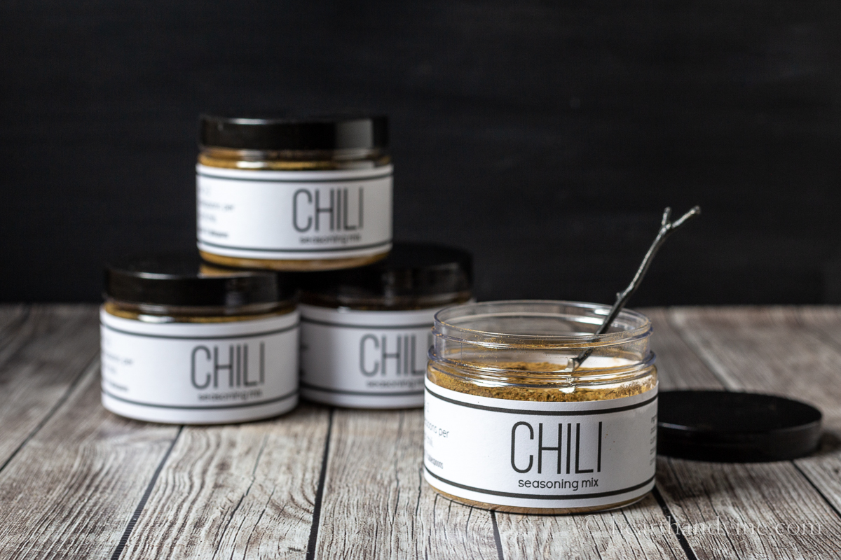 Four jars of chili seasoning mix. Three are stacked and one is opened with a small spoon inside.