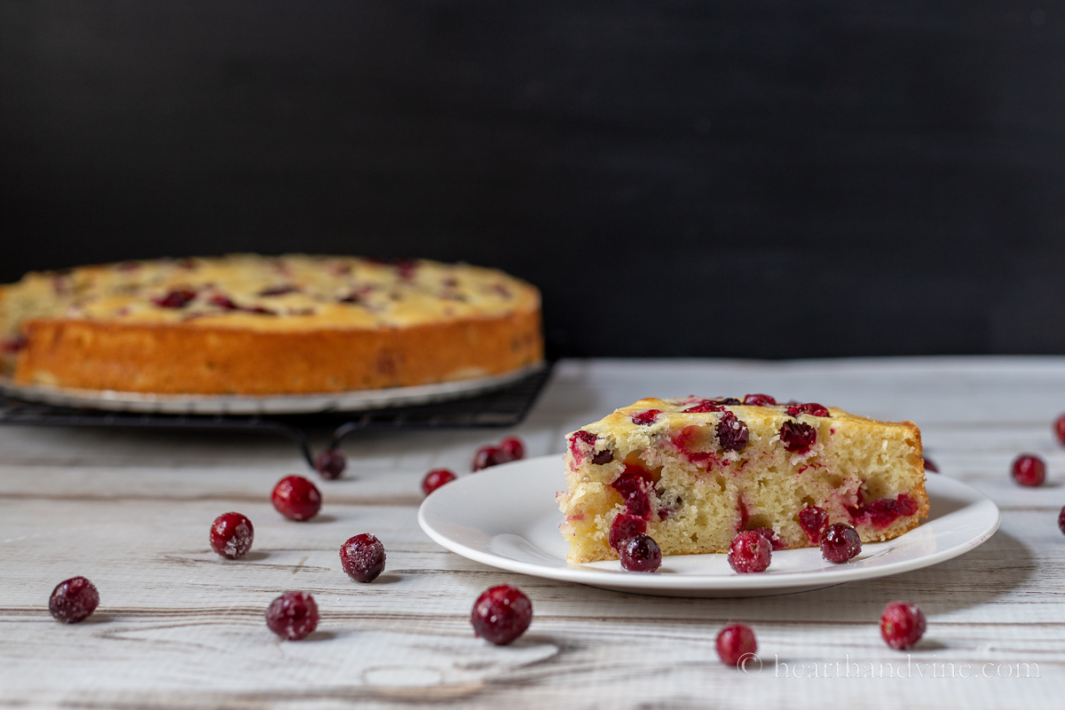 A slice of cranberry cake on a white plate with sugared cranberries scattered around. The remaining cake in the background.