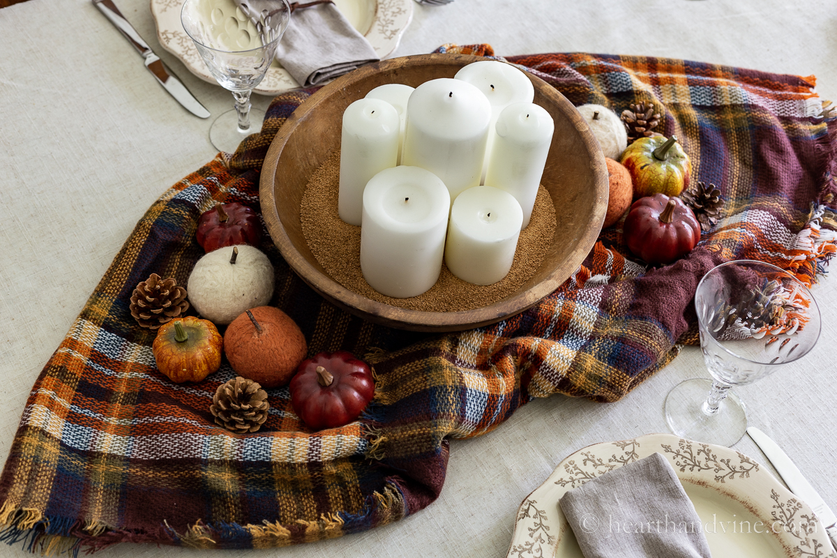 Dough bowl centerpiece on a fall plaid scarf on a table with pumpkins, and pine cones.