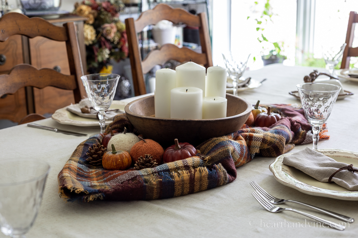 Fall table decorated with a round wooden dough bowl centerpiece with pillar candles on plaid scarf in autumnal colors.