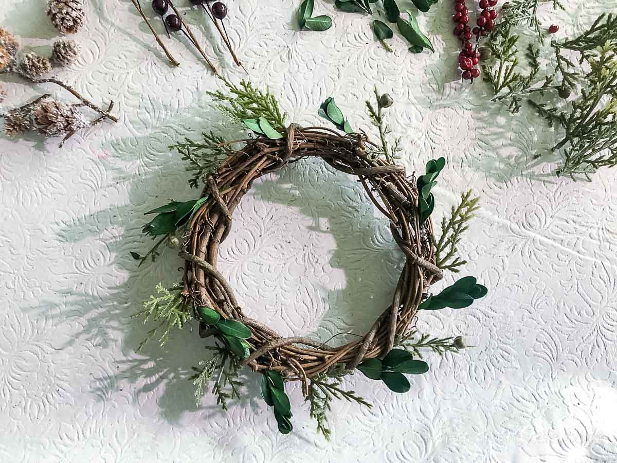 Six inch grapevine wreath with pieces of faux juniper and boxwood glued around the edge.