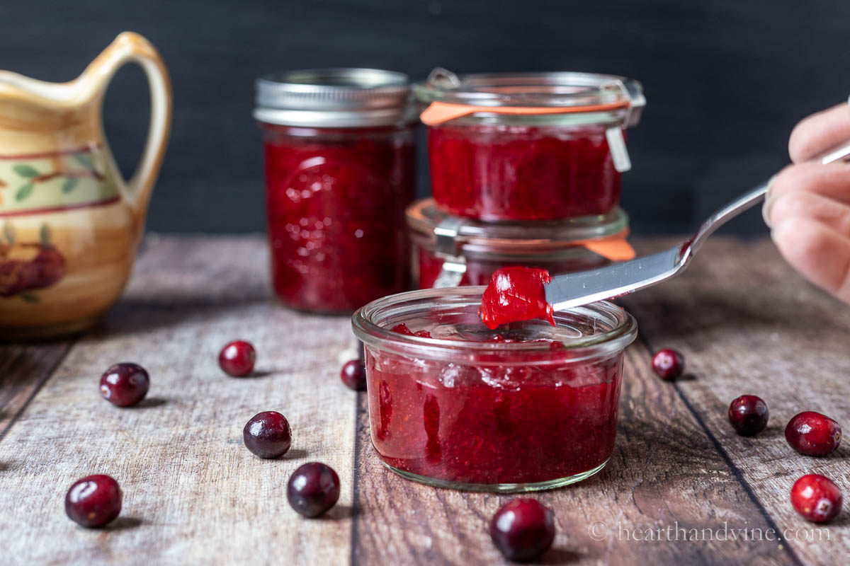A mini weck jar with cranberry jam and a knife lifting up a bit of jam. Fresh cranberries and more jars around the table.