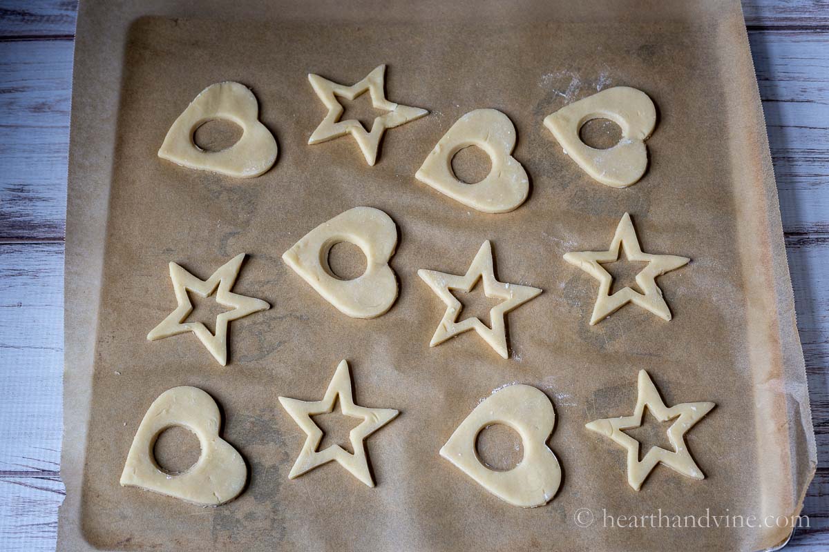 Sugar cookies cut out in stars and hearts with a circle or star cut out in the middle on parchment paper.