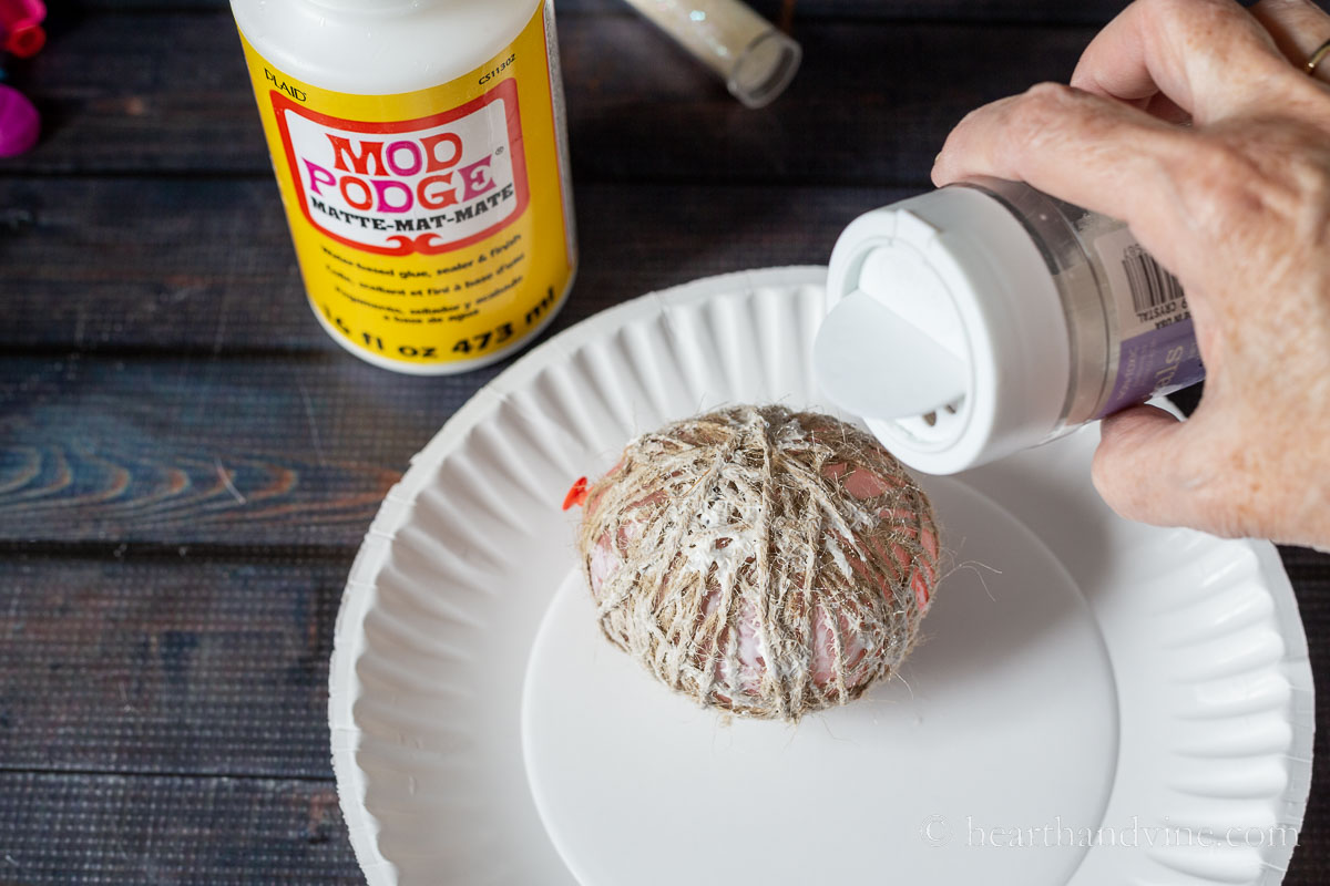 A small balloon covered with twine and glue on a plate. A shaker of glitter over the ball.