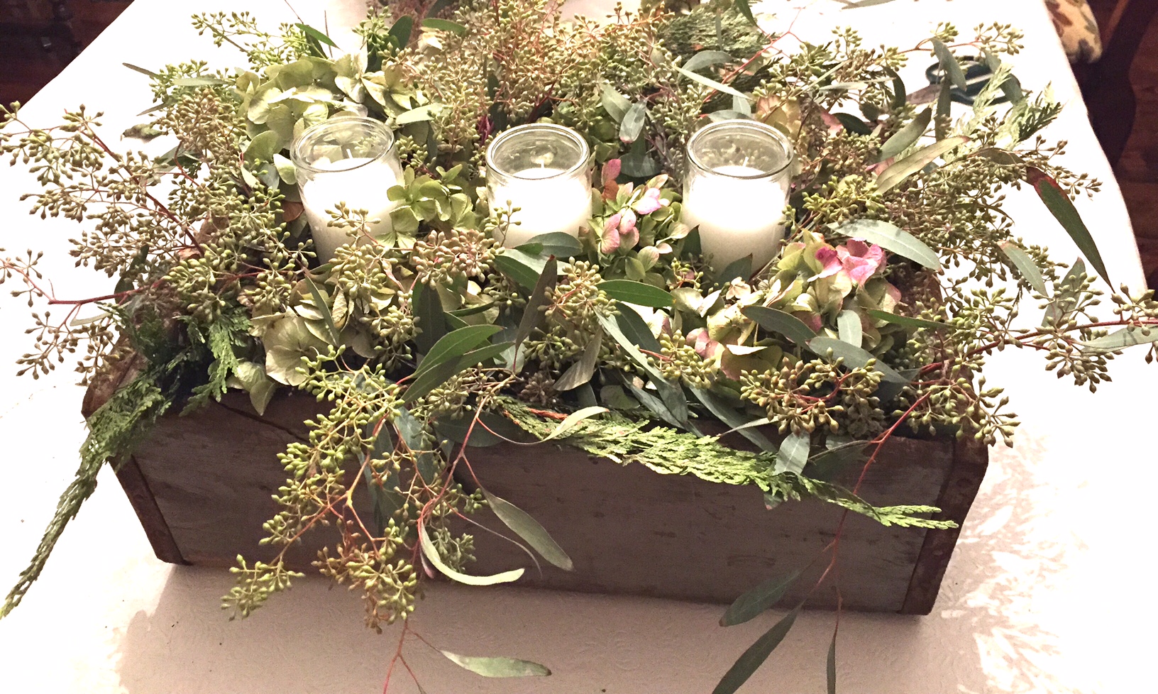 Antique box with an arrangement of dried hydrangea flowers, candles in glass, cedar branches and seeded eucalyptus.