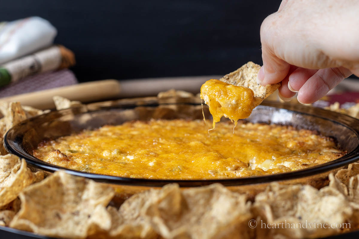 A hand lifting a bit of corn dip on a scoop tortilla above a platter with corn dip and chips.