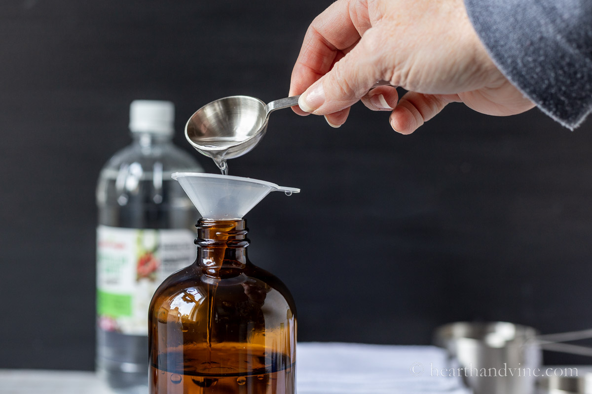 A hand pouring liquid from a measuring spoon into a funnel set in a glass amber bottle.