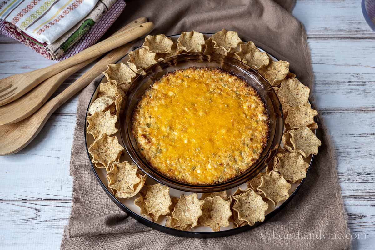 Baked hot corn dip with tortilla scoop chips around the side on a tan fabric next to wooden kitchen spoons.