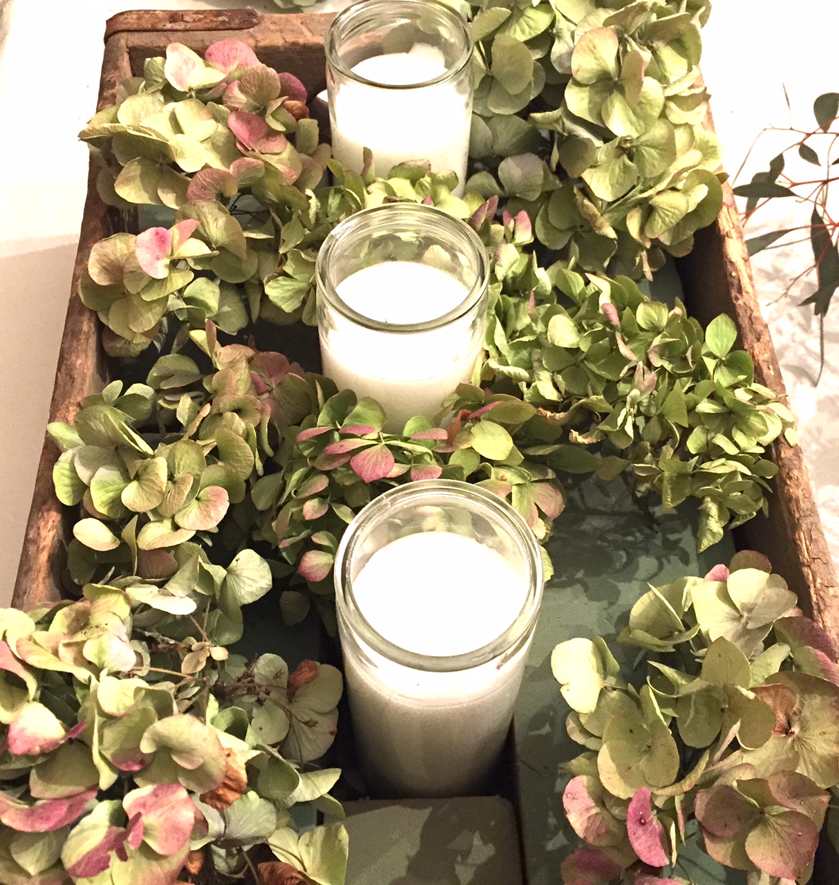Large wooden box with floral foam, three glass candles and dried hydrangea flowers.