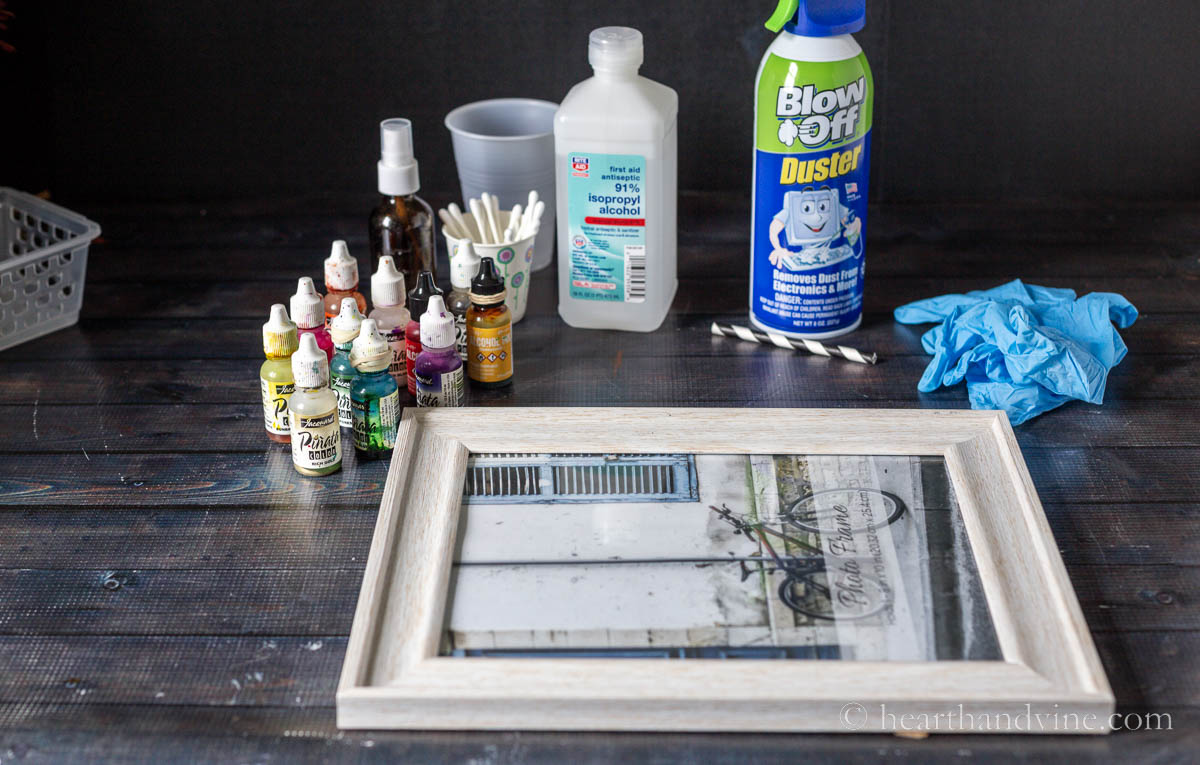 Supplies for painting on a photo frame with alcohol inks, including inks, isopropyl alcohol, canned air, gloves and cotton swabs.