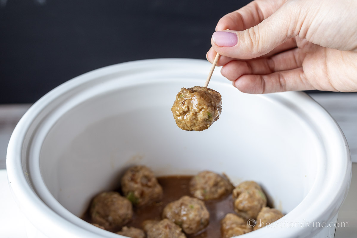 A hand holding a meatloaf meatball with a toothpick above a slow cooker of the same meatballs.