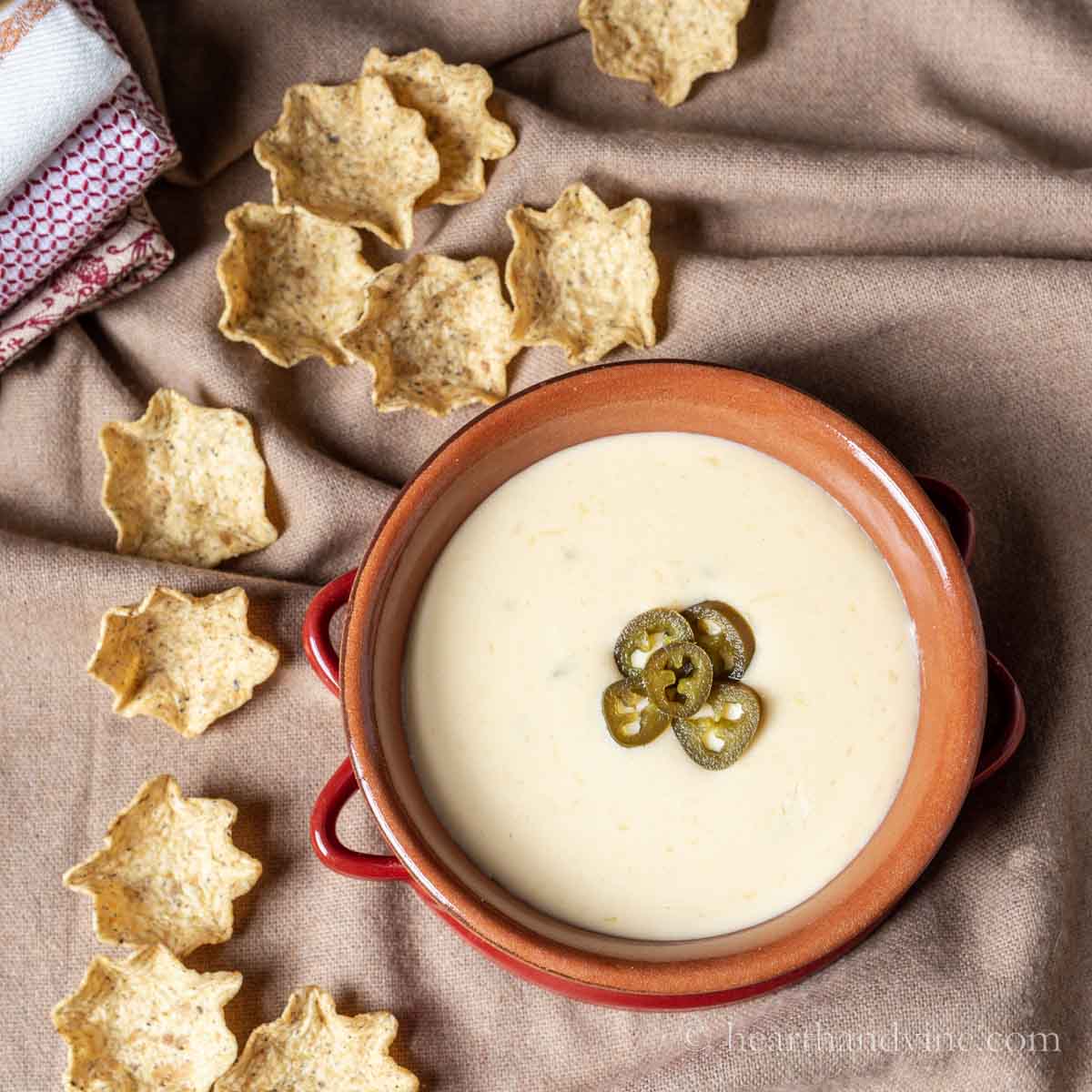 Bowl of queso dip with jalapeno slices in the middle and tortilla chips around the left side.