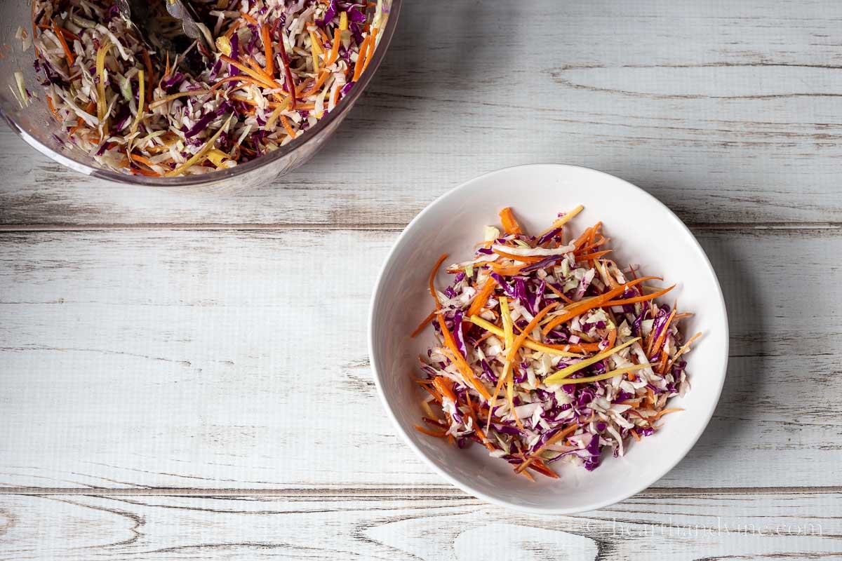 A white bowl filled with rainbow slaw near a large clear bowl of same salad.
