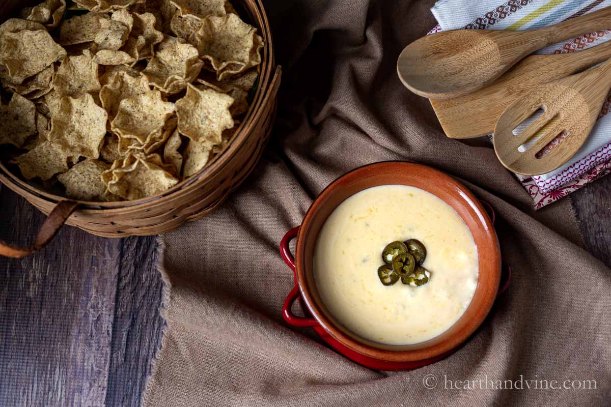 Bowl of queso dip next to a basket of tortilla chips and some wooden kitchen spoons.