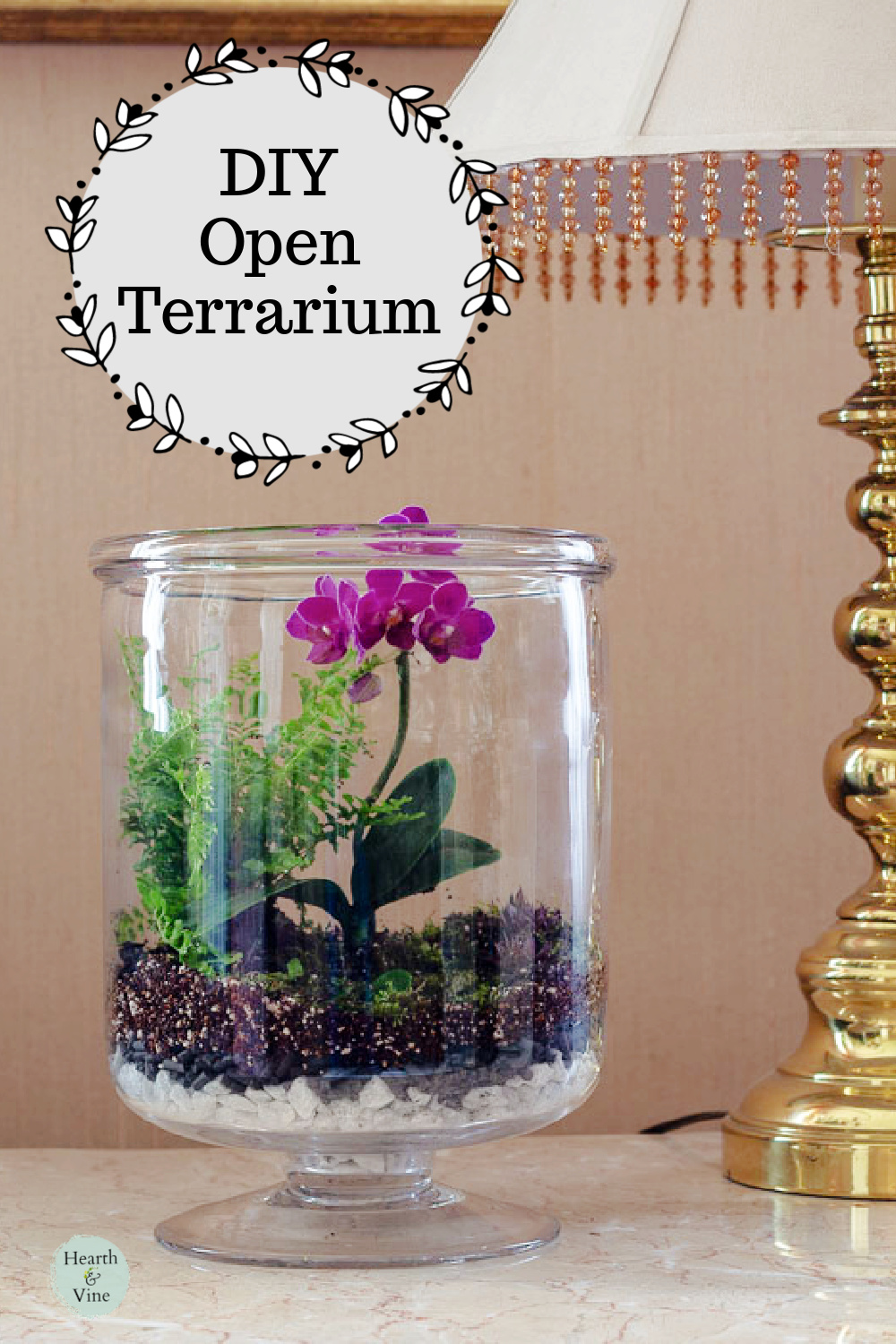 Open terrarium with a fern and pink orchid on a table next to a lamp.