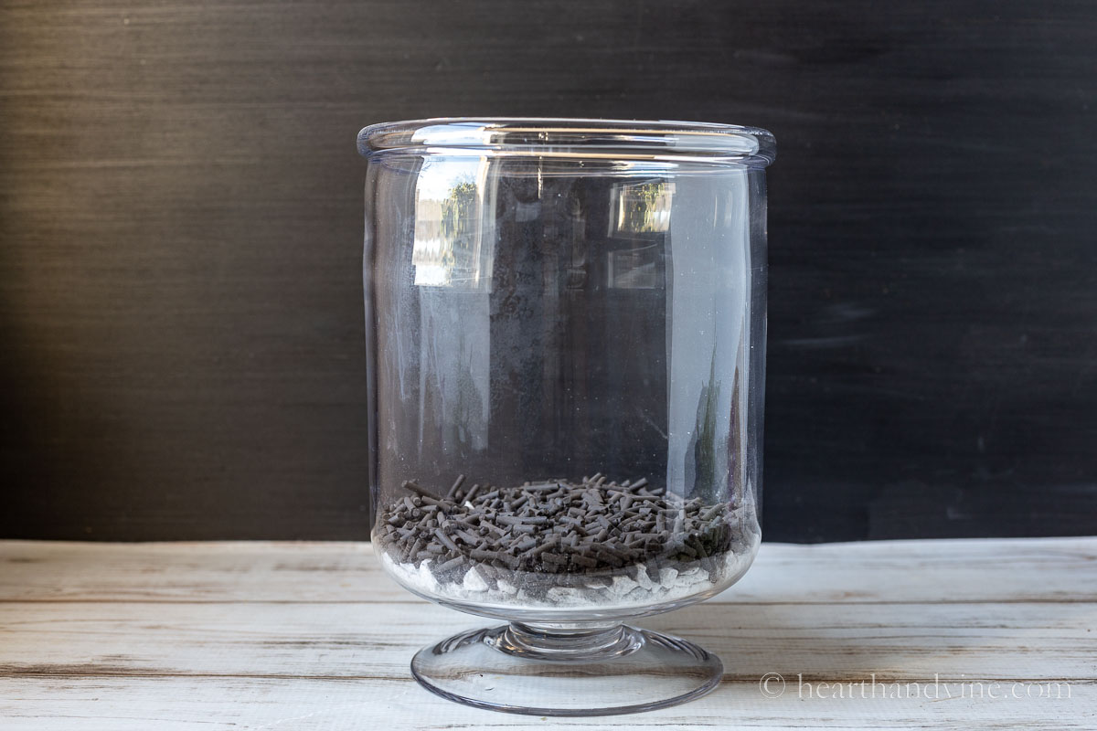 Tall open glass pedestal vase with a layer of white rocks followed by a layer of activated charcoal pellets.
