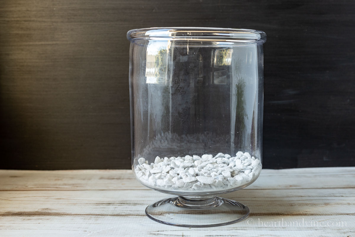 Tall glass vase on a pedestal with a layer of white gravel in the bottom.