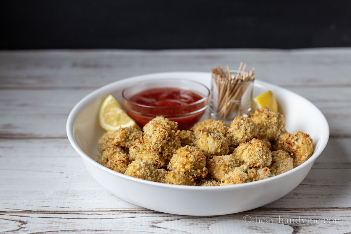 Side view of serving bowl with lemon wedges, a mini bowl of cocktails sauce, a cup with toothpicks and the baked crab balls.