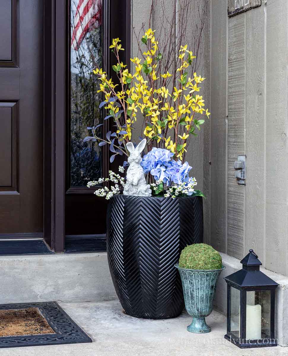 Right side of front porch with a black planter filled with artificial flowers and a cement bunny. below a small urn with a moss ball and an electric lantern.
