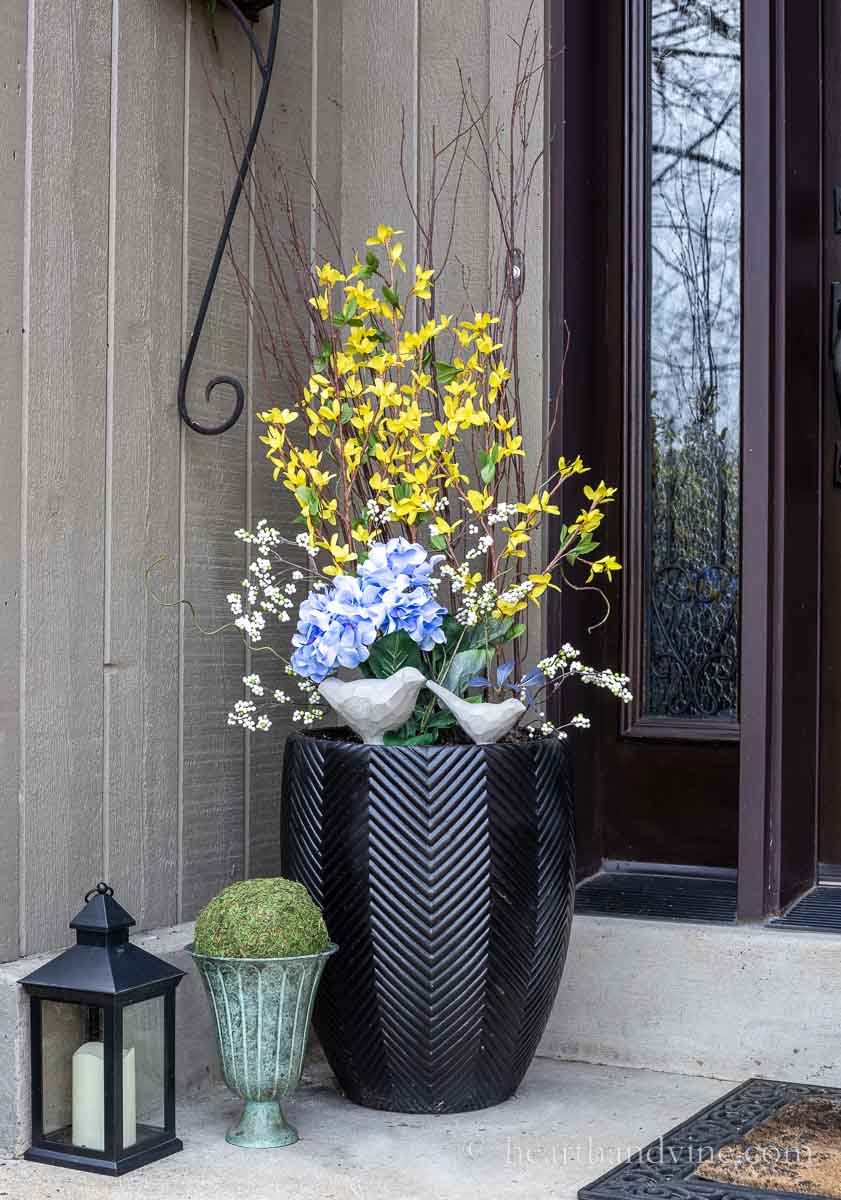 Left side of front door with a planter filled with faux forsythia, blue flowers and white berries. Also two cement birds.