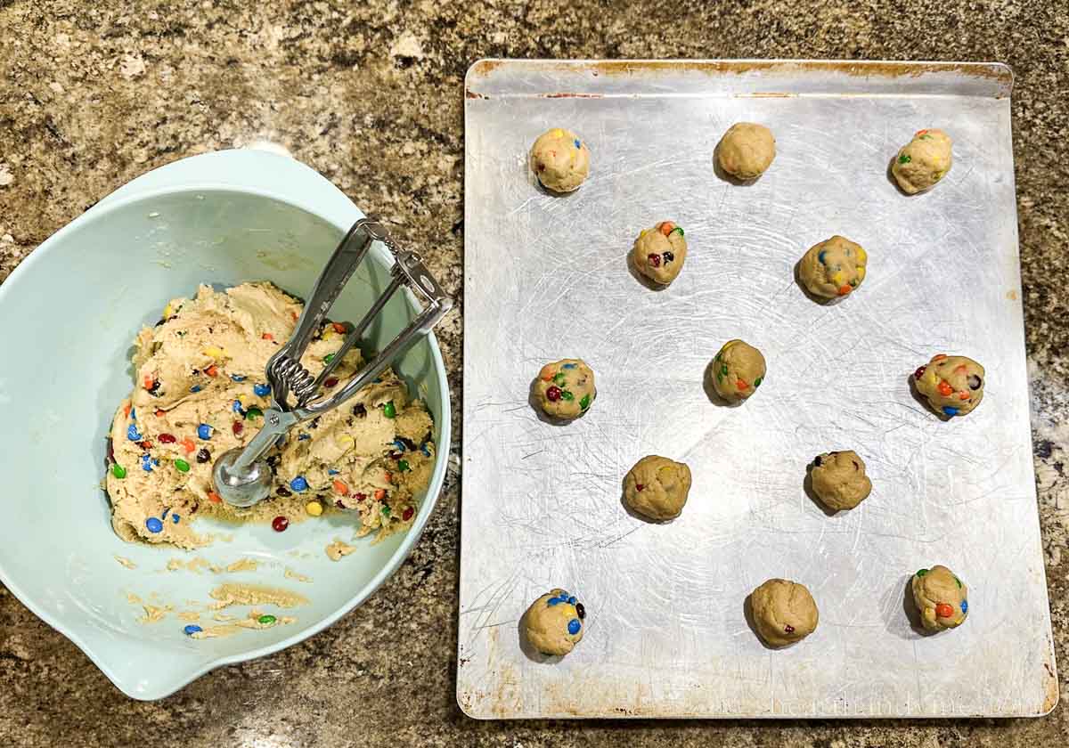 Cookie dough in a mixing bowl with a small cookie scoop next to a baking sheet with cookie dough balls.