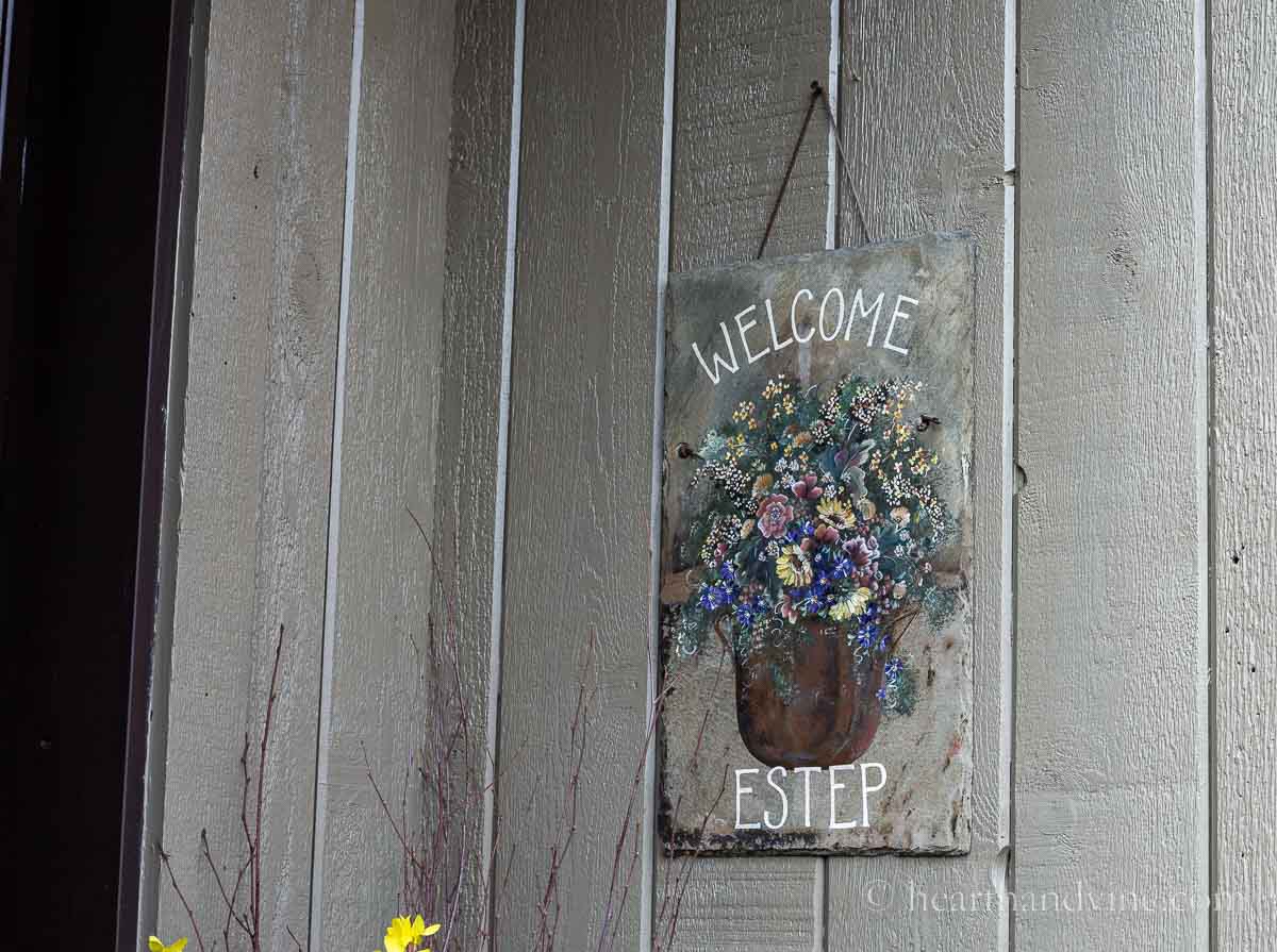 A slate hanging painted with a pot of flowers and the words Welcome and Estep on it.