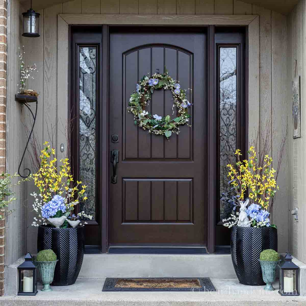 Front porch decorated for spring with faux flowers in planters, a garland wreath, lanterns and moss balls.