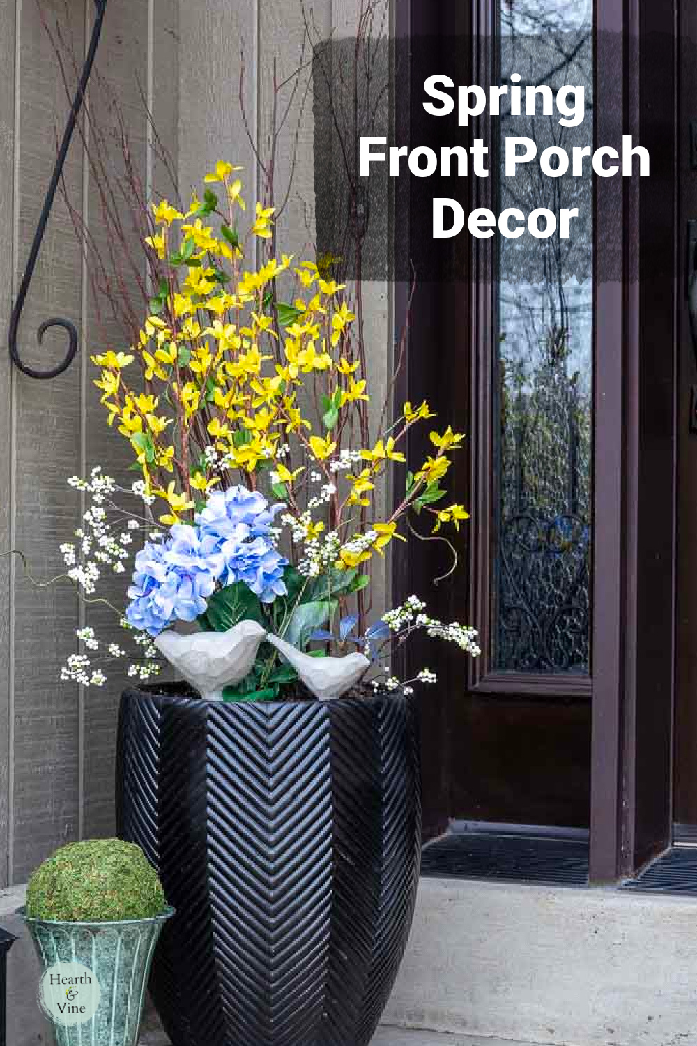 Black planter on front porch with faux blue and yellow flower and two cement birds.