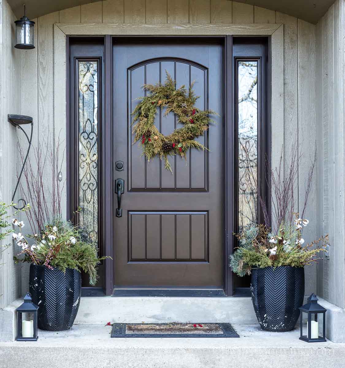 Front porch decorated for winter at the end of the season. A juniper wreath with berries and pinecones, two planters with more greenery and faux cotton picks and two fake candle lanterns.